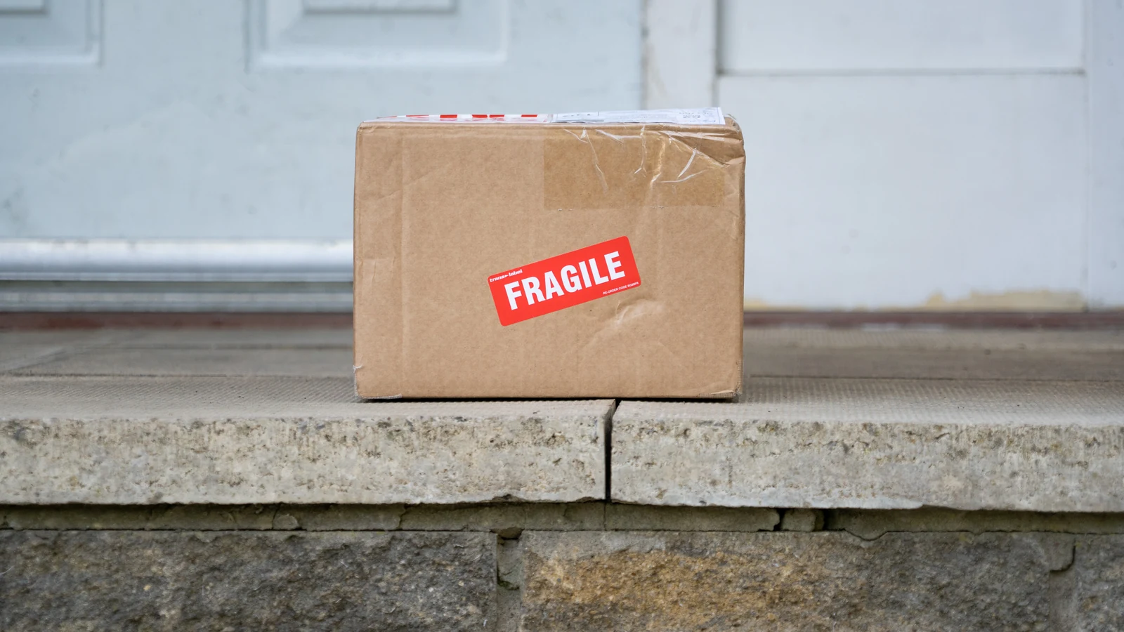 Помятая картонная коробка с красной наклейкой Fragile стоит на каменном крыльце