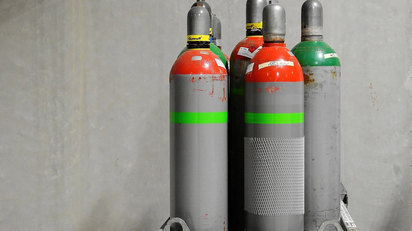 Стальные газовые баллоны с цветными горловинами стоят на подставке у стены склада
